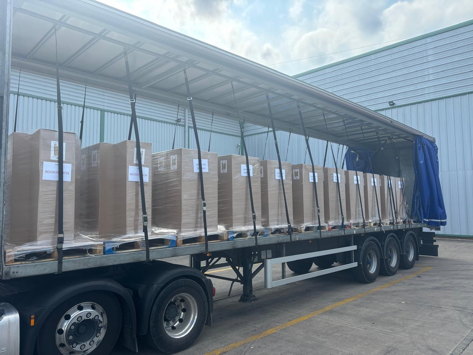 Open truck trailer loaded with large wrapped pallets standing outside a warehouse.